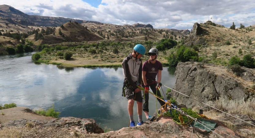 Two people wearing safety gear are secured by ropes near the edge of a cliff. One person appears to be an instructor, giving direction to the other person. There is a lake below them.